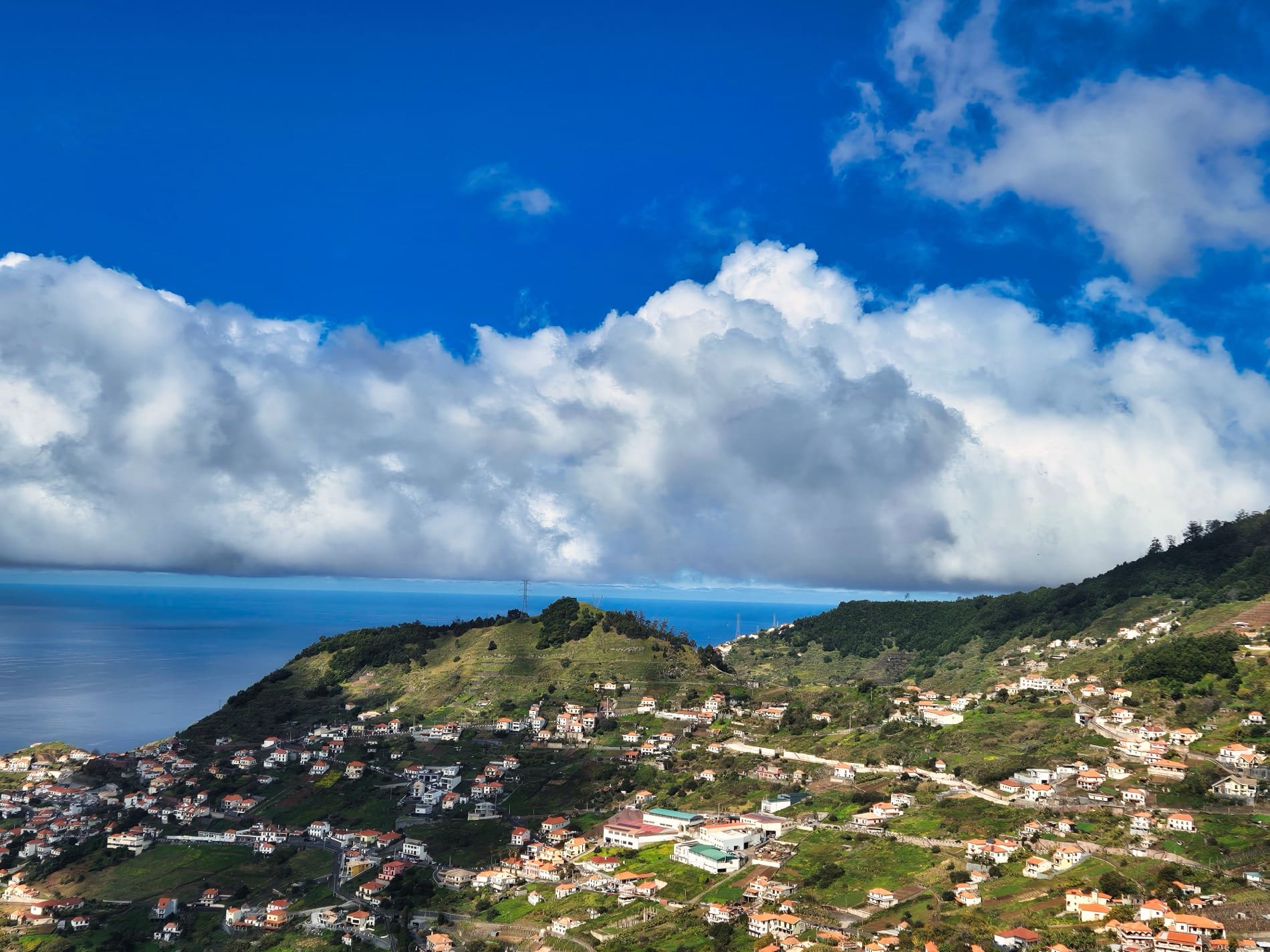 Panoramic view of coastal village nestled on green hills overlooking the ocean, with dramatic clouds above.